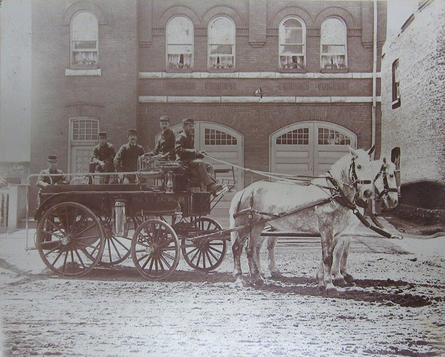 Vintage sepia photograph circa 1900. Orillia Fire Brigade in front of brick Fire Hall. Five men sit atop an industrious fire carriage, lead by two white horses over sandy road.