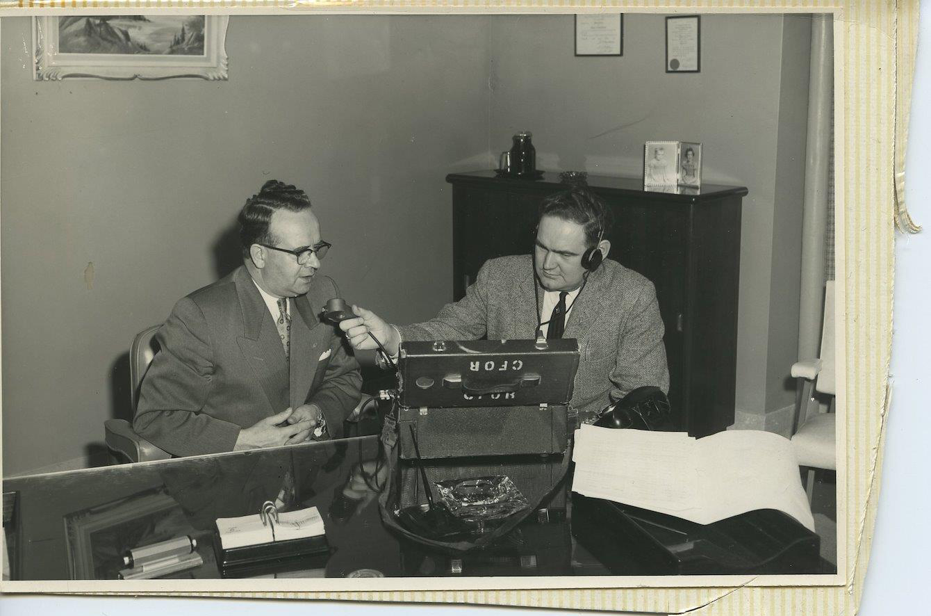 Vintage black and white photograph with torn, yellowed edges circa 1950. Pete McGarvey sits at large desk with CFOR recording box and holds microphone to his left towards a well-dressed man in black-rimmed glasses.