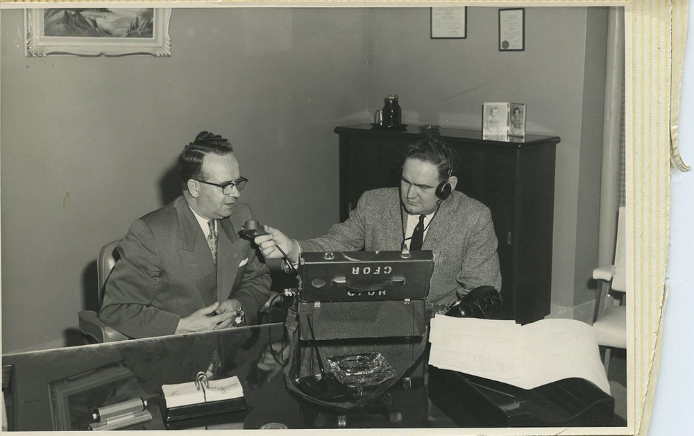 Vintage black and white photograph with torn, yellowed edges circa 1950. Pete McGarvey sits at large desk with CFOR recording box and holds microphone to his left towards a well-dressed man in black-rimmed glasses.