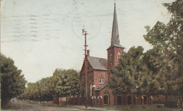 Colour-tinted postcard of a rural Orillia steeple church built with red brick, along a densely tree-lined street, circa 1909.