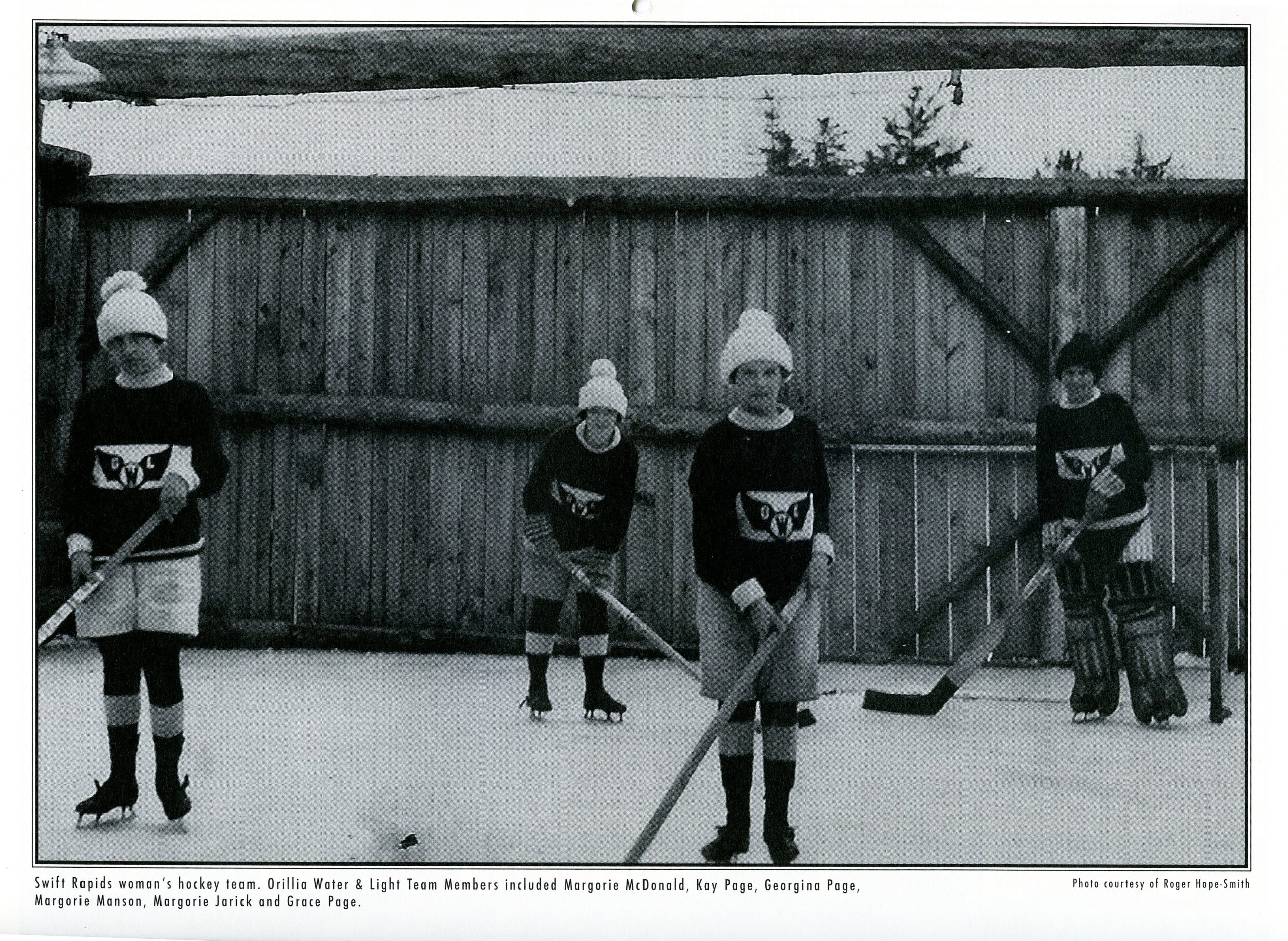 Vintage black and white photograph circa 1925. Four young women dressed warmly in uniform pose in front of a tall wooden fence holding hockey sticks over an outdoor ice rink. Additional details are printed below image.