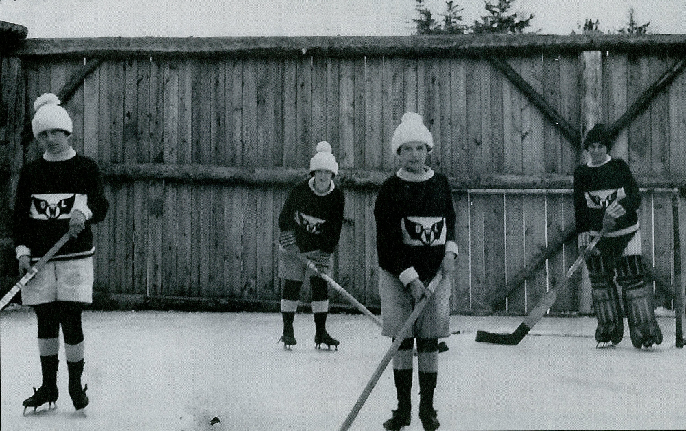 Vintage black and white photograph circa 1925. Four young women dressed warmly in uniform pose in front of a tall wooden fence holding hockey sticks over an outdoor ice rink. Additional details are printed below image.