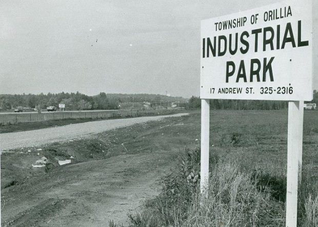 Vintage black and white photograph circa 1968. Large, white, wooden road sign in the countryside reads, Township of Orillia, Industrial Park, 17 Andrew St. 325-2316