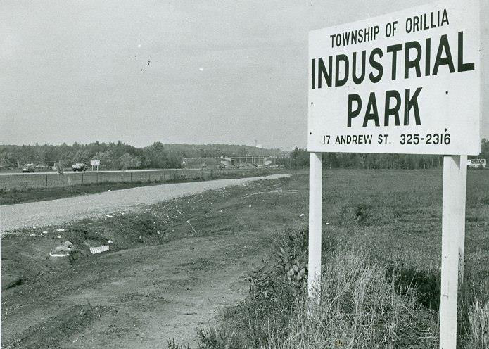 Vintage black and white photograph circa 1968. Large, white, wooden road sign in the countryside reads, "Township of Orillia, Industrial Park, 17 Andrew St. 325-2316"