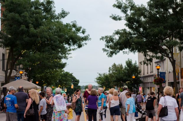 Warm, August evening in 2016. Smiling patrons converse on a crowded street downtown Orillia, canopied by green trees and twinkle lights, lined with glowing street lamps and historic buildings.