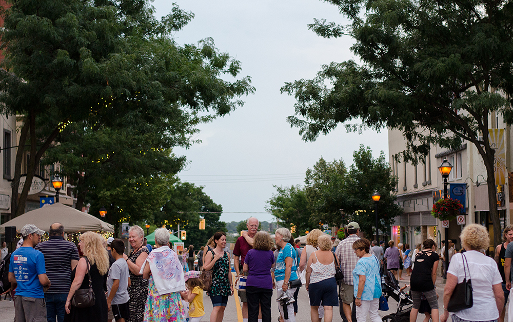 Warm, August evening in 2016. Smiling patrons converse on a crowded street downtown Orillia, canopied by green trees and twinkle lights, lined with glowing street lamps and historic buildings.