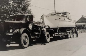 Black and white photograph of a white yacht, placed on a trailer, attached to a black truck. F-X Lachance is standing to the left of the truck. “The Notre-Dame, 1952. belonging to the Curé Bélanger of Berthier”