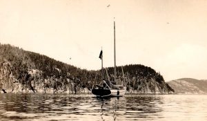 Black and white photograph of a small two-masted sailboat, sails unfurled, moored in a few centimetres of water. The title 