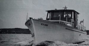 Black and white photograph of a small yacht seen from the front, sailing on the river. A man wearing a hat is at the controls in the cabin. A Canadian flag flies at the back. 