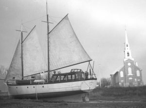 Black and white photograph of a two-masted sailboat, sails unfurled, on the beach near the church of Saint-Laurent. 