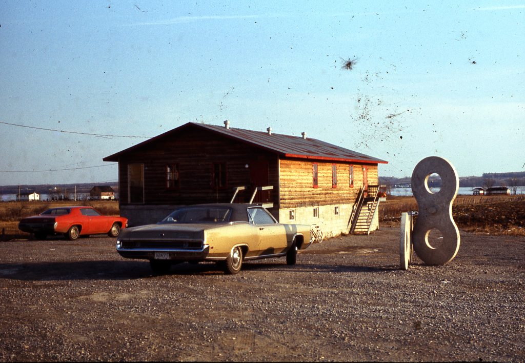 Wooden building next to which is a sculpture in the shape of an “8”. Two cars are parked in front. The St. Lawrence River can be seen in the distance.