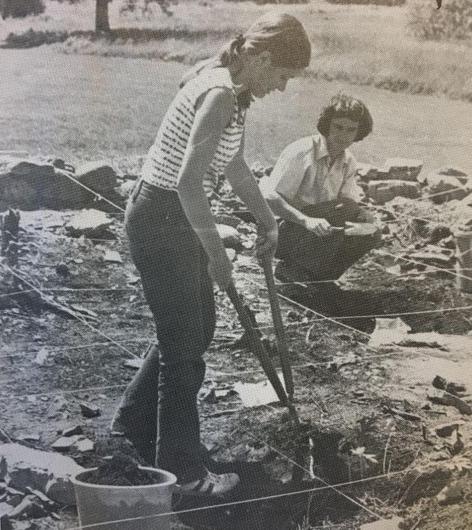 Black-and-white photo of a standing woman holding long scissors and a young man crouching behind, doing archaeological excavations in a rope cadrilateral.