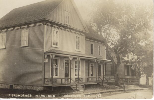 Black and white photo of a two-storey house with a gallery. On the first floor is a general store.