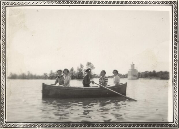 Black and white photo of five young adults in a rowboat. In the background, a small lighthouse in the water.