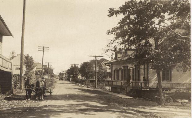 Black and white photo of a dirt street, with houses on either side. On the left, two horses harnessed to a cart and on the right, an old-fashioned carriage.