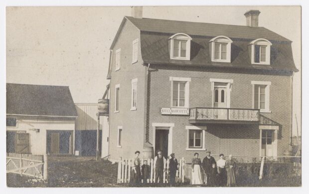 Black and white photo of nine people in front of a three-story brick house. Above the door is the inscription: “Hôtel Marcotte”.