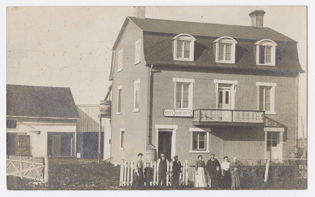 Black and white photo of nine people in front of a three-story brick house. Above the door is the inscription: “Hôtel Marcotte”.