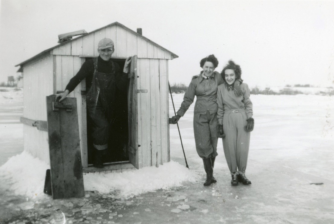 Black and white photo of two women and a man next to an ice fishing shack.