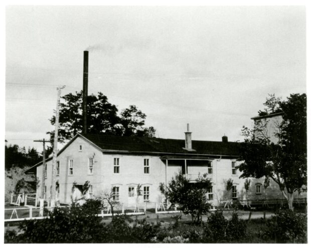 Black and white photo of a long two-storey building with a tall chimney.