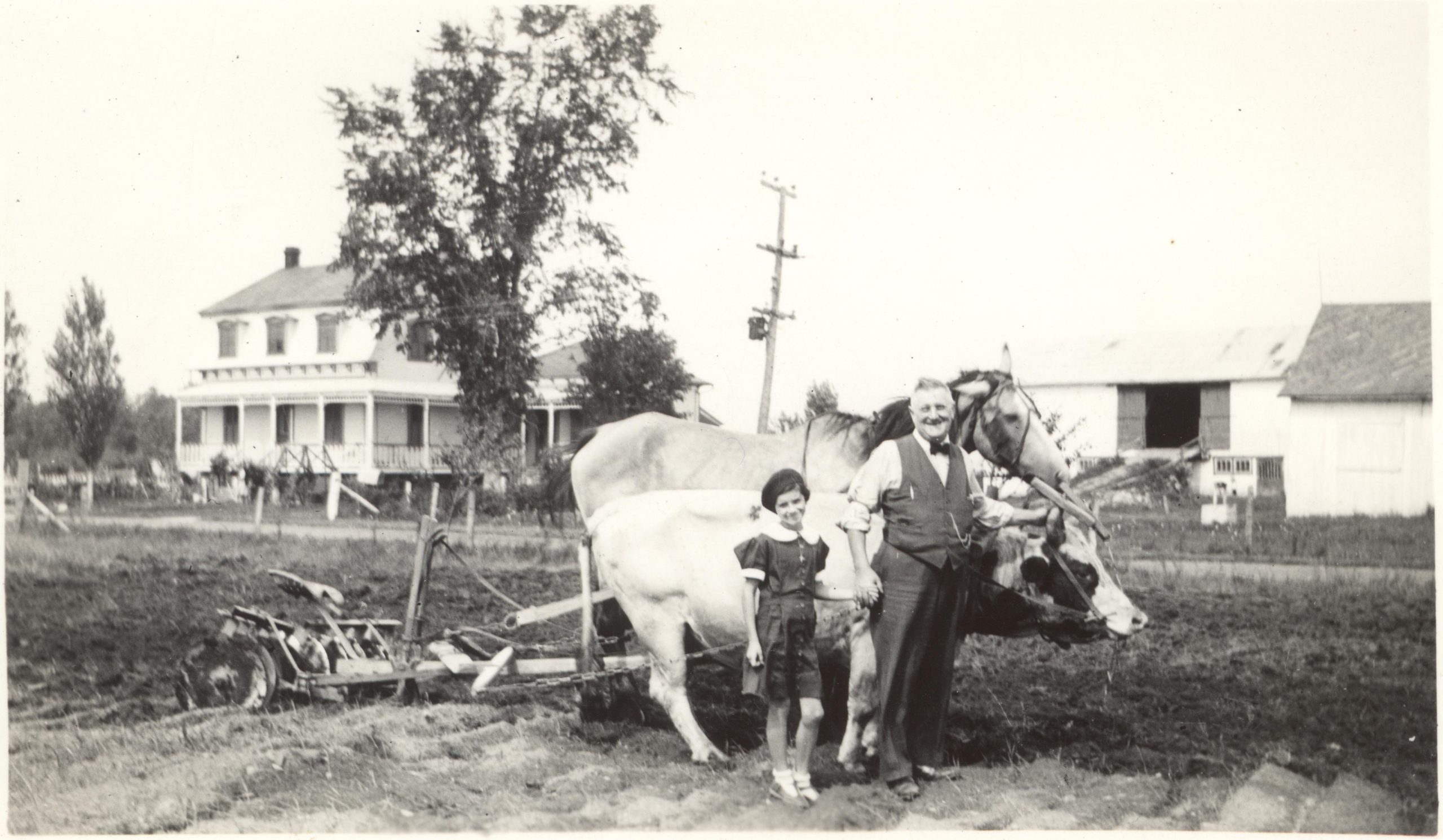 Black and white photograph of a man and child beside a harnessed ox and horse. Behind, a two-storey house and farm buildings.