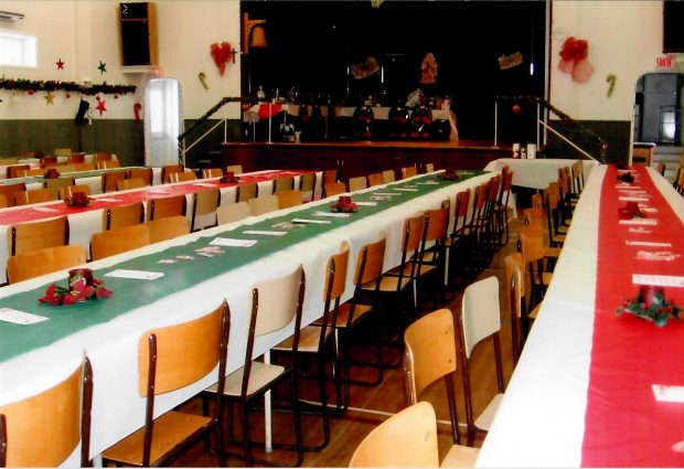 Room decorated for Christmas with five large tables and wooden chairs. In the background, a small theatre stage.