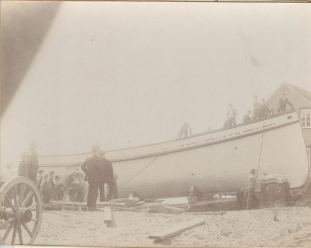 Black and white photo of a ship under construction. Several men are beside and in the ship.