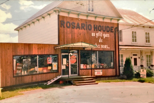 Photo of a large two-storey house with two buildings. The building on the left is occupied by a general store. The storefront reads “Rosario Houde - un p'tit gars de chez nous...”.