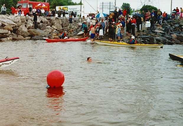 A group of people and two kayakers waiting for a swimmer to arrive on the edge of a wharf. In the background, an emergency vehicle.