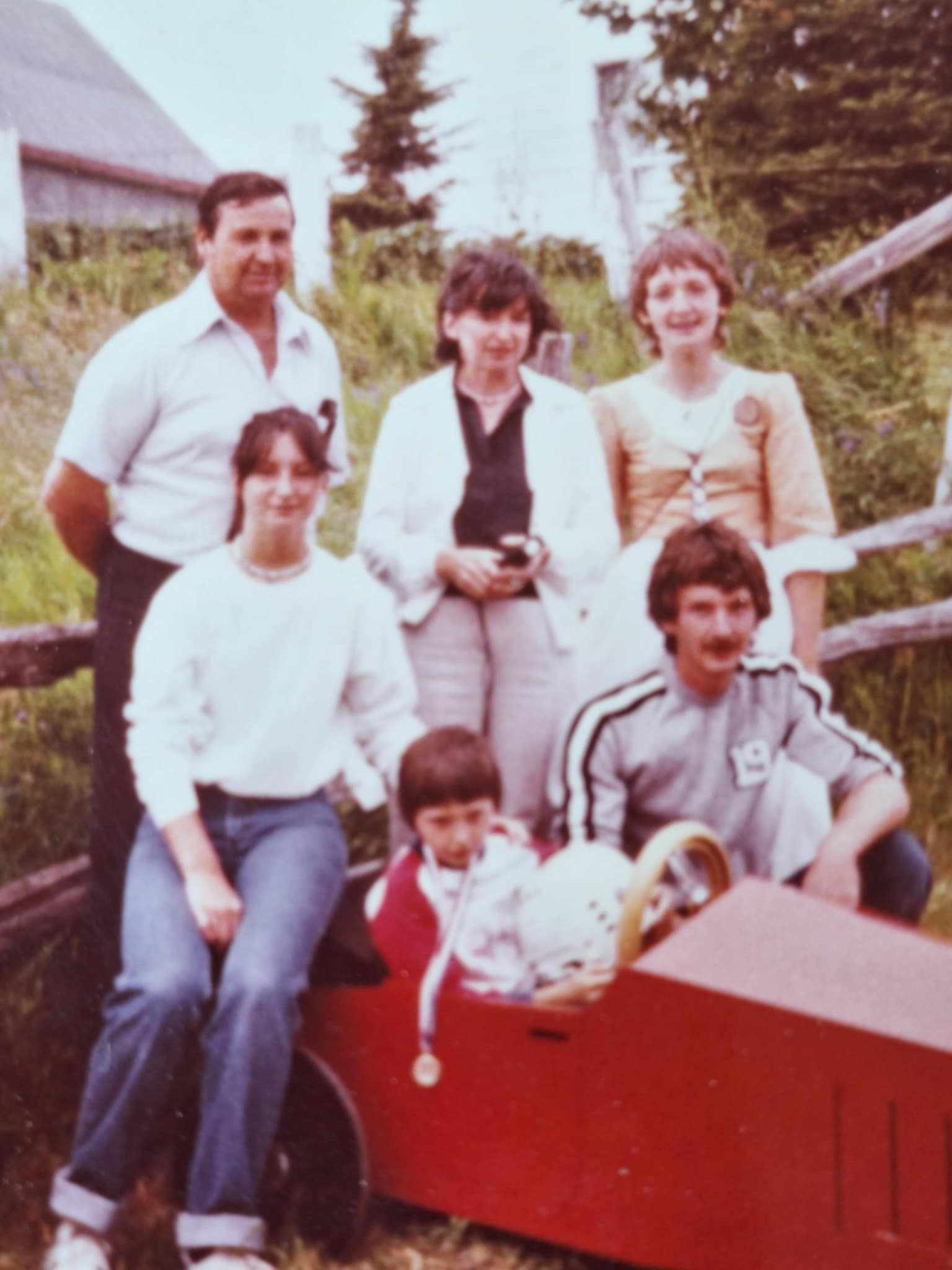 Five adults pose with a child seated in a small wooden racing car.