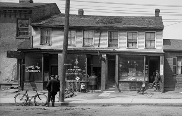 Black and white historical photo of a two-story wooden building with signs and a bicycle parked outside and two men standing by the laundry shop