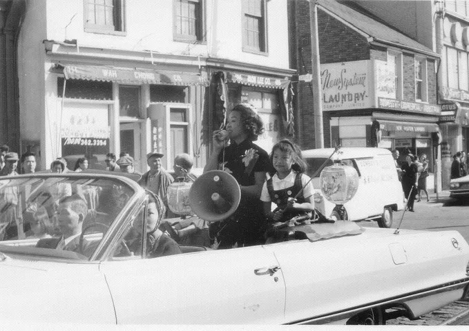 Black and white photo from the 1960s showing Jean Lumb speaking through a megaphone in a parade, with a child beside her in a convertible car, and a sign in the background