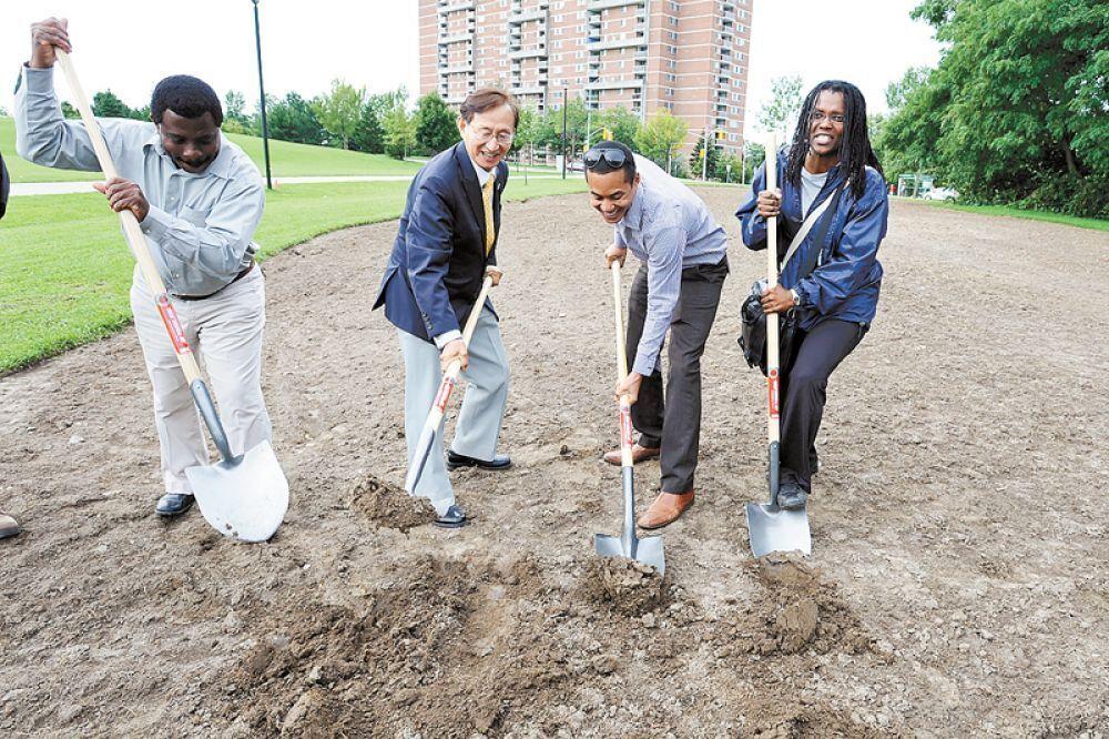 Image shows four people with shovels at a groundbreaking ceremony. They are smiling and digging into the earth, with a grassy field and high-rise buildings in the background.