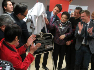 A group of people in a room, including members of the Wong Association and Justin Trudeau, clapping while unveiling a commemorative plaque.