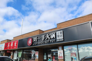 A one story red brick building with a black sign, red logo and white Chinese characters and English letters reading Liuyishou Hot Pot over glass windows with promotional signs on them.