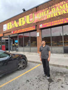 A man standing in front of a restaurant named 'BABU,' which advertises all-day breakfast and lunch combos and now features Chef Damu's signature biryani.