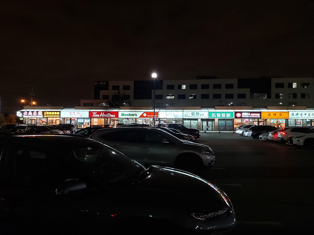 A night view of a well-lit one storey shopping plaza with various storefronts, including Tim Hortons and Beckers, among others with Asian characters on their signs.