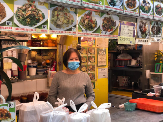 A smiling woman with a mask standing behind the counter in a busy food stall, surrounded by pictures of dishes and take-out bags, with a health inspection pass sign visible.