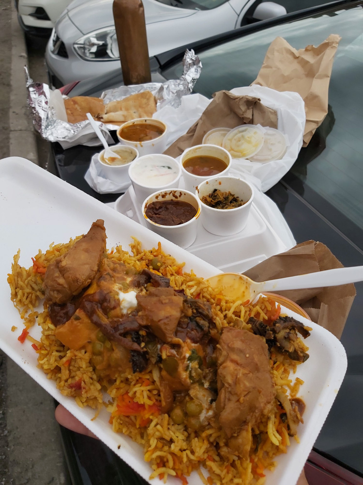 A close-up of a plate filled with biryani and various side dishes, served in a take-out container, with more food wrapped in aluminum foil in the background, placed on the hood of a parked car.