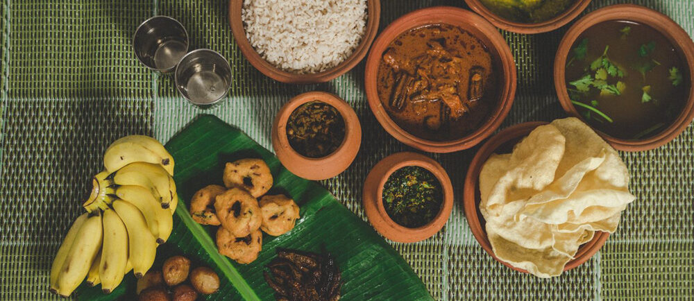 A spread of various traditional dishes on a table with rice, curries, and bananas on a green woven mat. The food is served in earthenware pots.