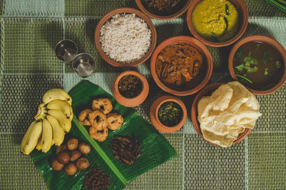 A spread of various traditional dishes on a table with rice, curries, and bananas on a green woven mat. The food is served in earthenware pots.