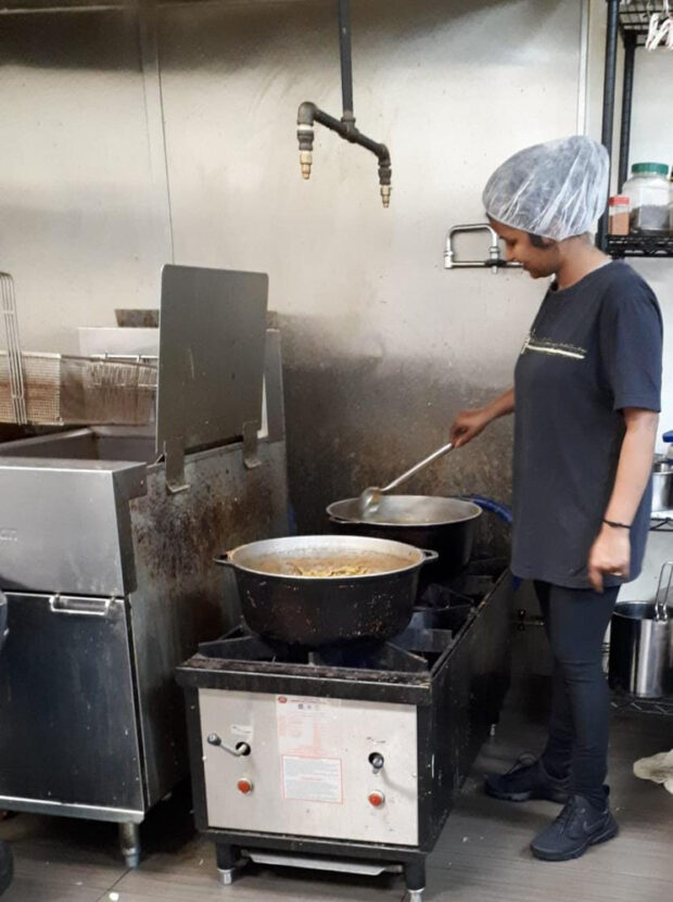 A woman in a kitchen, stirring a large pot on an industrial stove. She is wearing a hairnet and casual clothes, standing in a kitchen with metal surfaces.