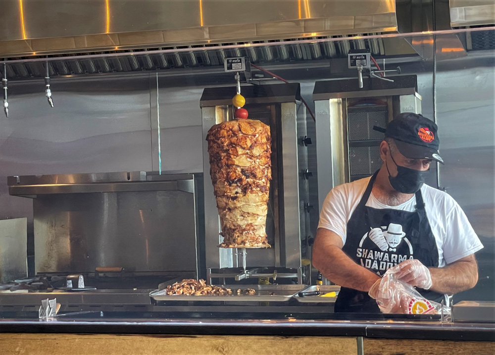 A man in a face mask and cap, wearing an apron reading 'Shawarma Daddy' cuts meat from a large rotisserie shawarma stand in a restaurant kitchen, with metal surfaces and cooking equipment around.