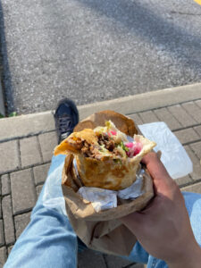 A person holding a shawarma wrap with meat, vegetables, and sauce visible. They are outdoors, with a paved surface of the parking lot beneath him.