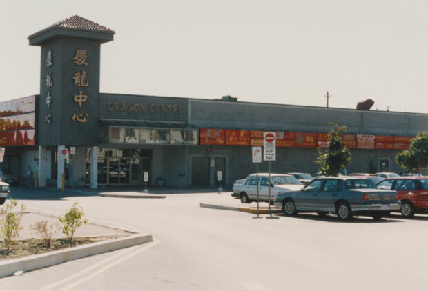 One story grey brick building with storefront signs, a grey brick tower with stylized Chinese characters and parked cars in a large parking lot.