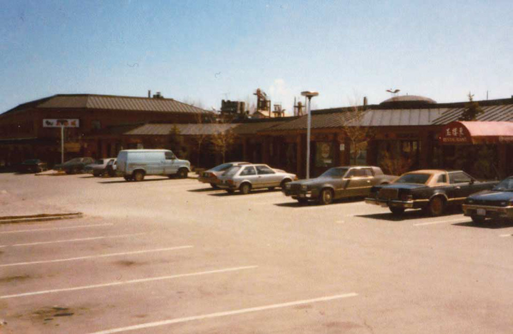 An analog photograph of a parking lot with parked cars in front of a one story row of restaurants and businesses part of Torchin Plaza in Agincourt.