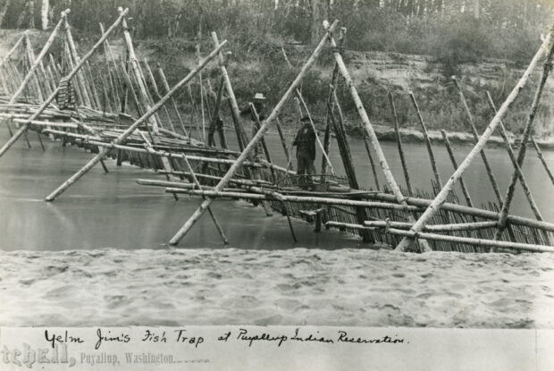 A black and white photograph of a fishing weir structure, made of long wooden sticks arranged in a triangle formation across a wooden bridge stretching across a river with two men standing on the bridge structure.