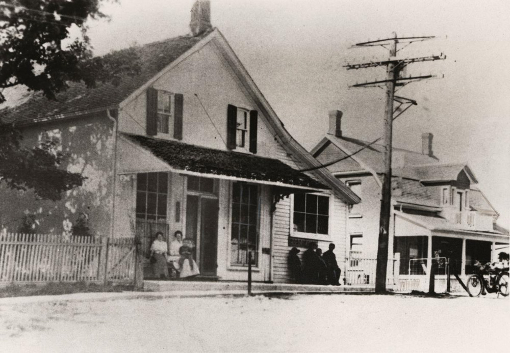 A black and white photograph featuring a two story white townhome with two small windows on the second floor, two large windows on the ground floor under a veranda, separated by a large door on a wide street with other homes, a picket fence, trees and a large power line. Seated in front of the windows are a woman and two young children to the left, and four dark figures to the right.