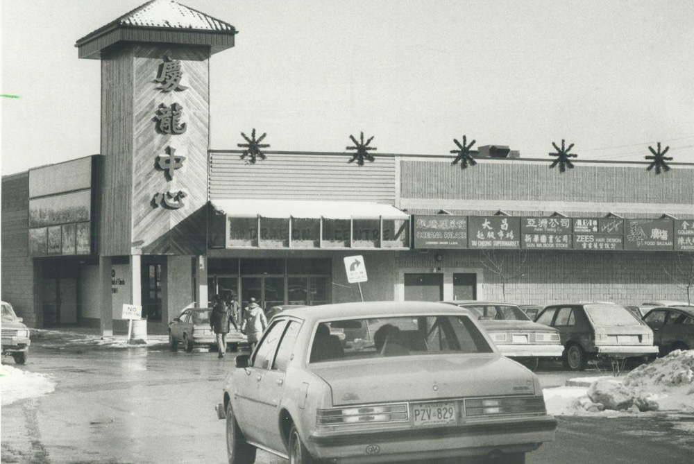 Black and white image of The Dragon Centre, then called The New Chinese Mall, with storefront signs, a tower with stylized Chinese characters and a full parking lot.