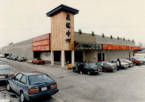 Analog photograph showing the Dragon Centre Mall with a grey brick facade, a wooden tower with stylized black Chinese characters, red storefront signs, and a full parking lot with patrons entering the mall.