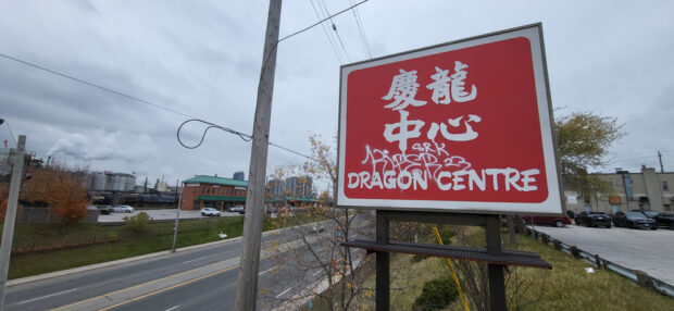 A coloured photograph of a large red sign atop a 6 lane road that reads Dragon Centre in English and Chinese characters.